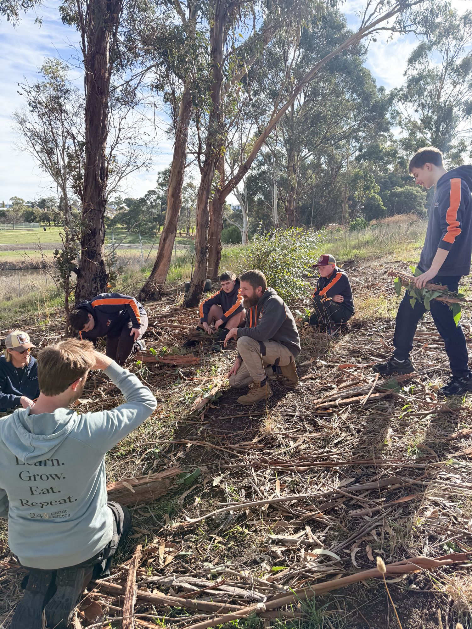 A group of people gathered under tall trees in an outdoor setting, crouching and sitting on the ground. They appear engaged in a discussion, conveying a sense of learning and teamwork. Sunlight filters through the leaves, creating a peaceful atmosphere.