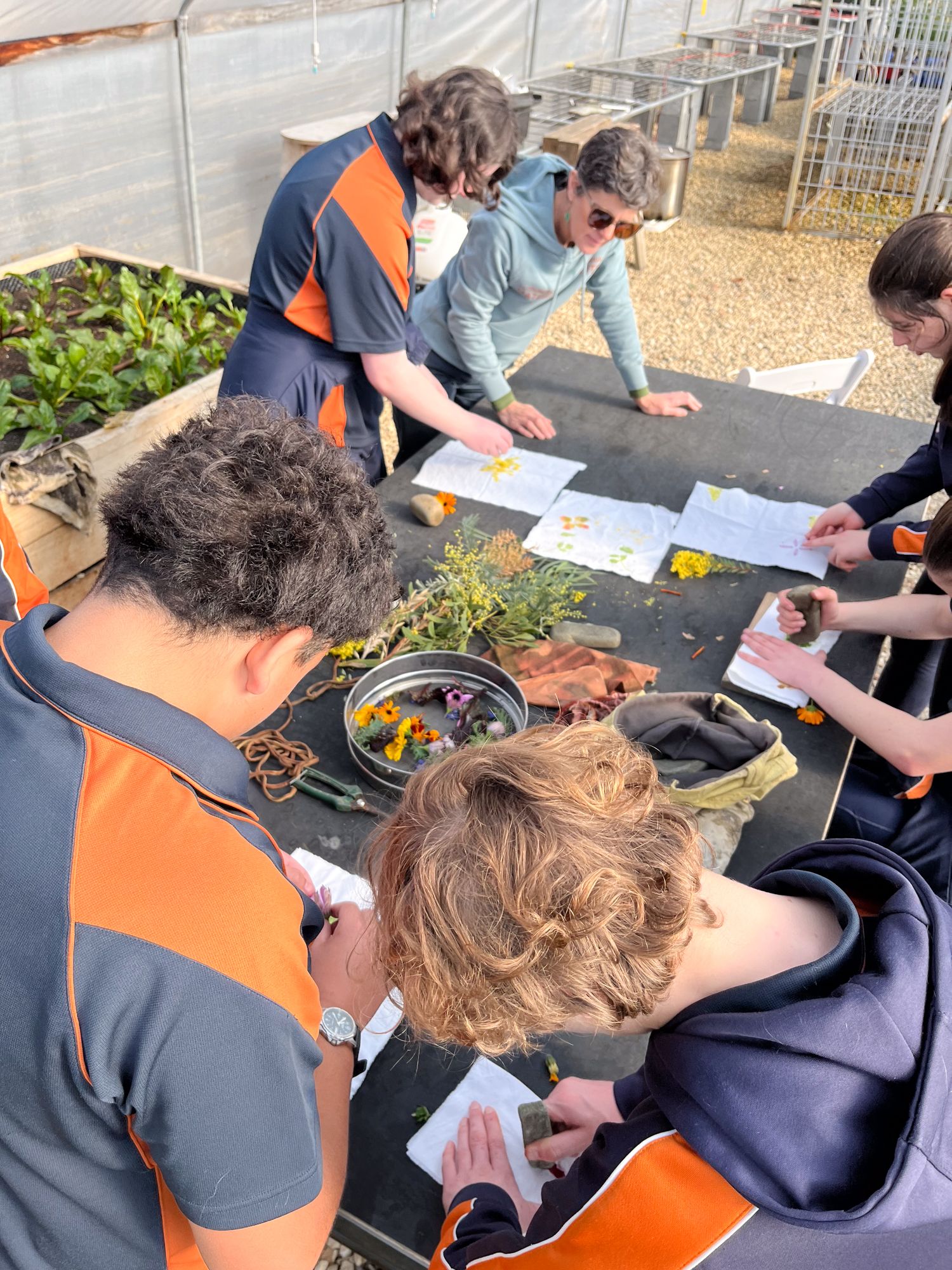 Students in uniforms and a teacher engage in a hands-on activity in a greenhouse. They're pressing flowers and leaves on fabric, creating a creative and educational atmosphere.