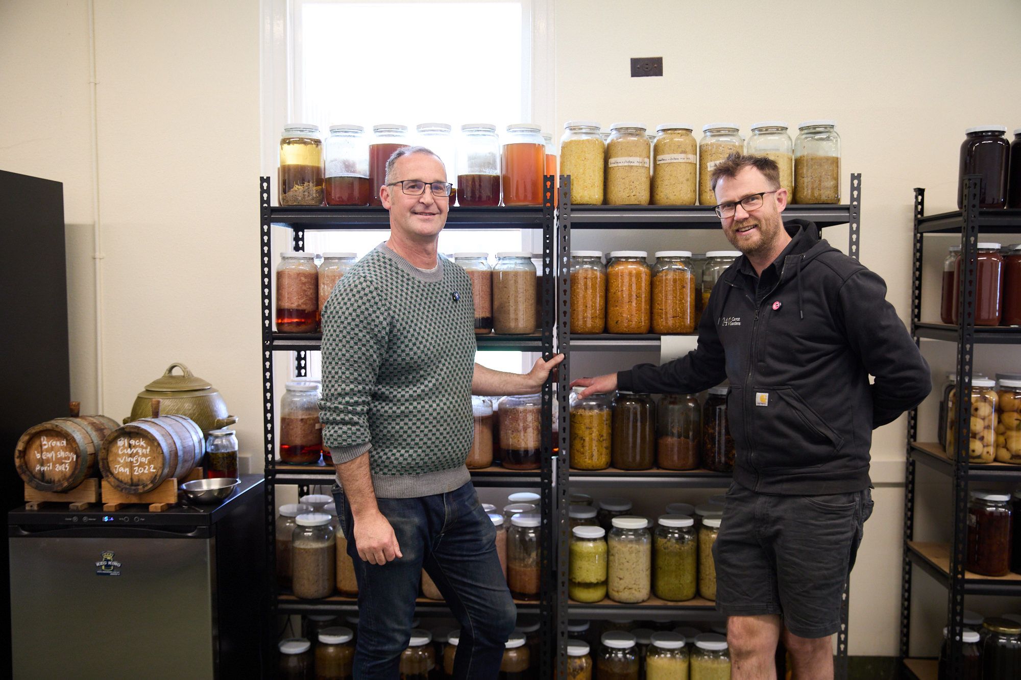Two men smiling in front of shelves filled with jars of pickled foods and fermented beverages. The room conveys a warm, inviting atmosphere.