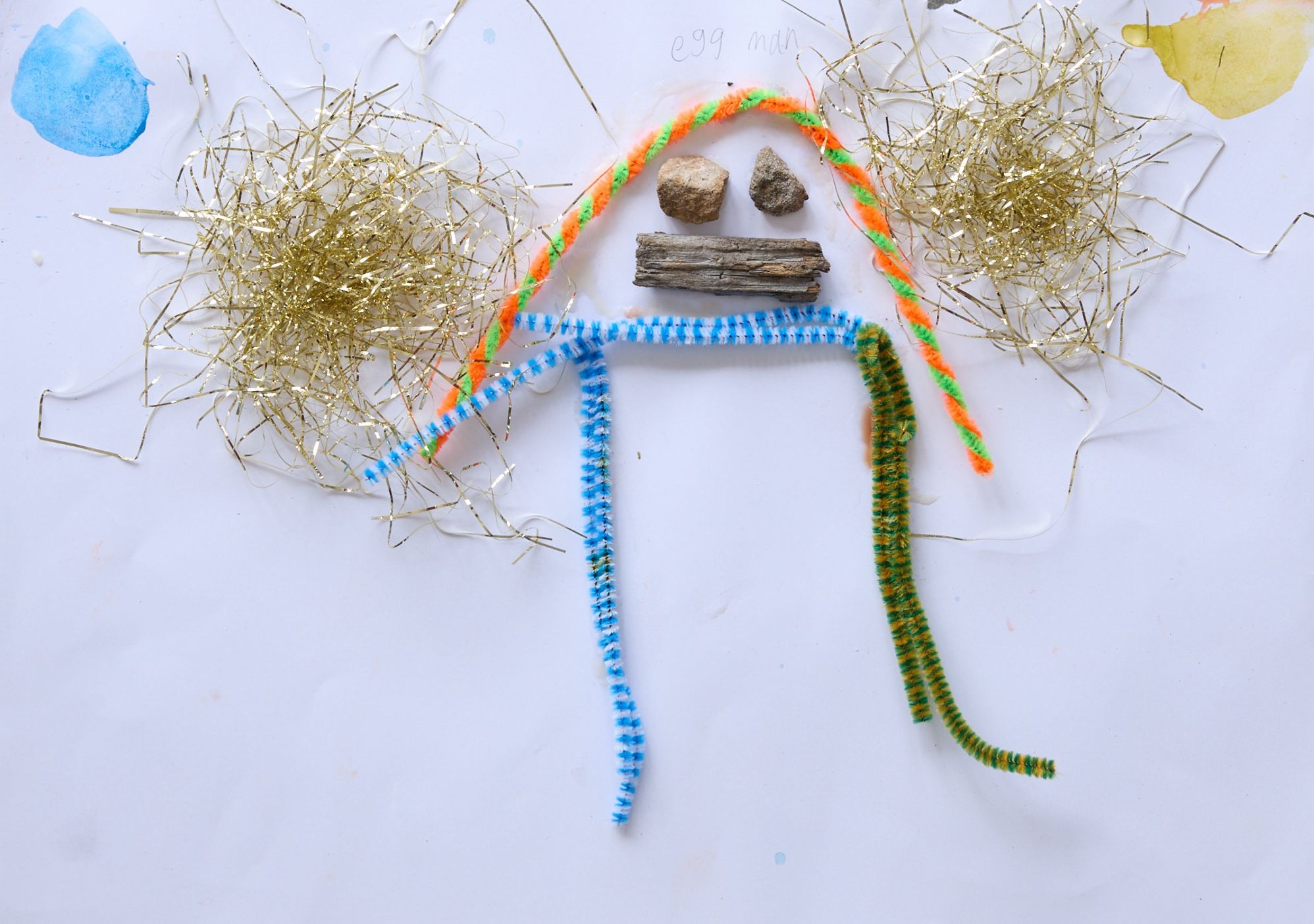 Colorful art piece on white paper with two glittery, gold yarn clusters, pipe cleaners in rainbow colors forming a stick figure. Pebbles and wood create facial features. Playful and whimsical tone.