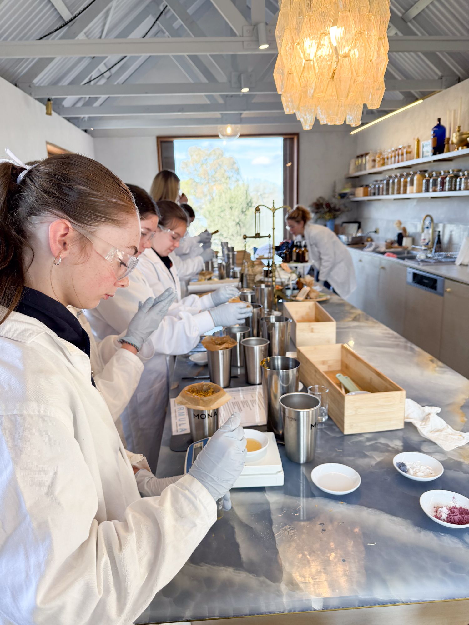 People in lab coats and goggles work in a bright, modern lab. They are focused on mixing ingredients at a long marble counter.