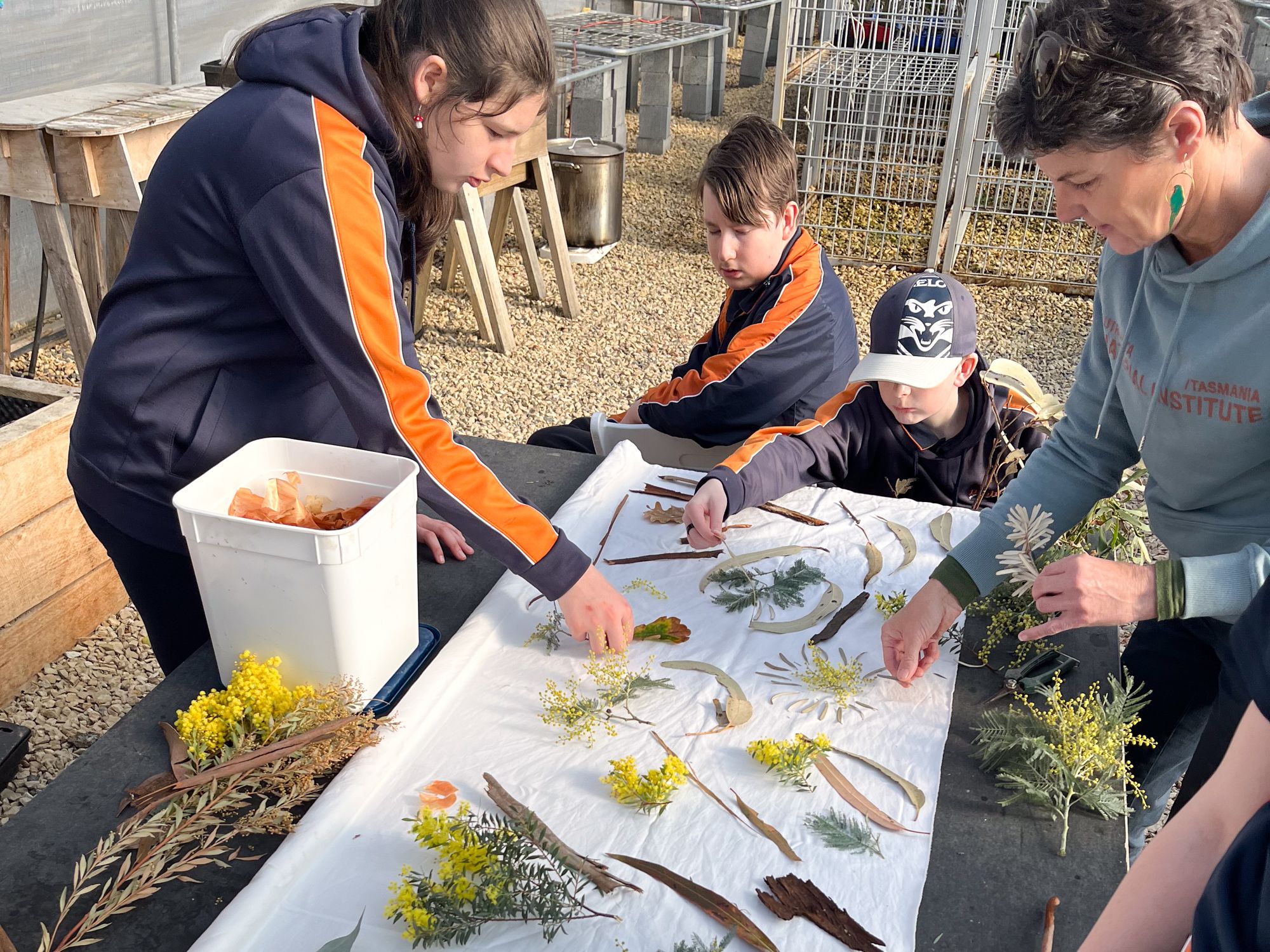 People in a greenhouse arrange leaves and flowers on white fabric. They wear blue uniforms and seem focused and collaborative in their activity.