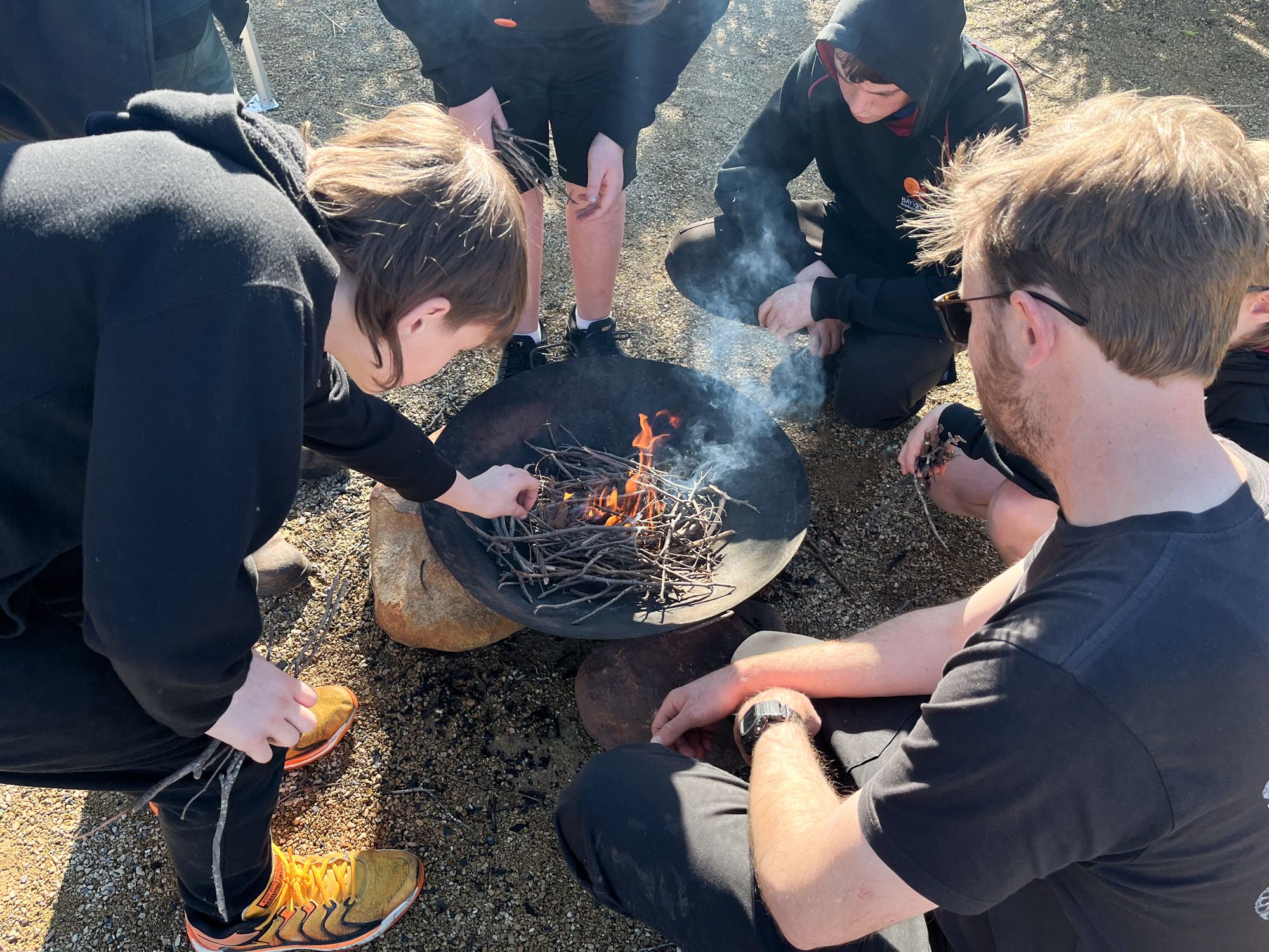 A group of people in black clothing sitting around and tending to a small campfire in a large metal bowl on gravel. The mood is focused and collaborative.