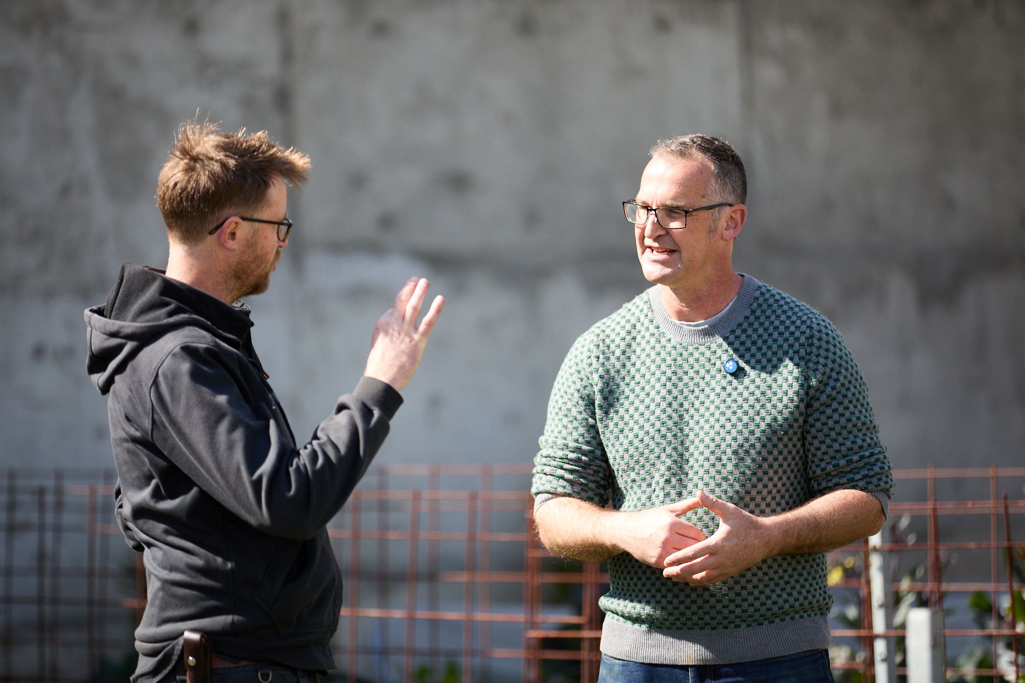 Two men having a friendly conversation outdoors. One in a dark hoodie uses hand gestures, while the other in a green sweater listens, both smiling.