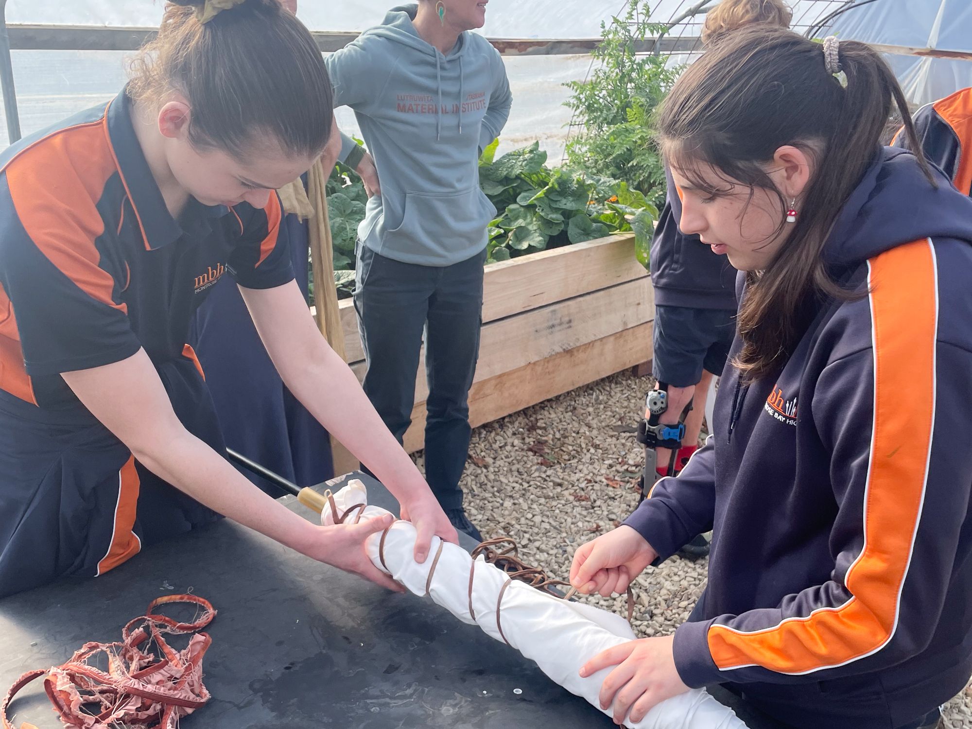 Two young women in uniforms work collaboratively to wrap a cylindrical object with twine in a greenhouse. A person in the background observes.