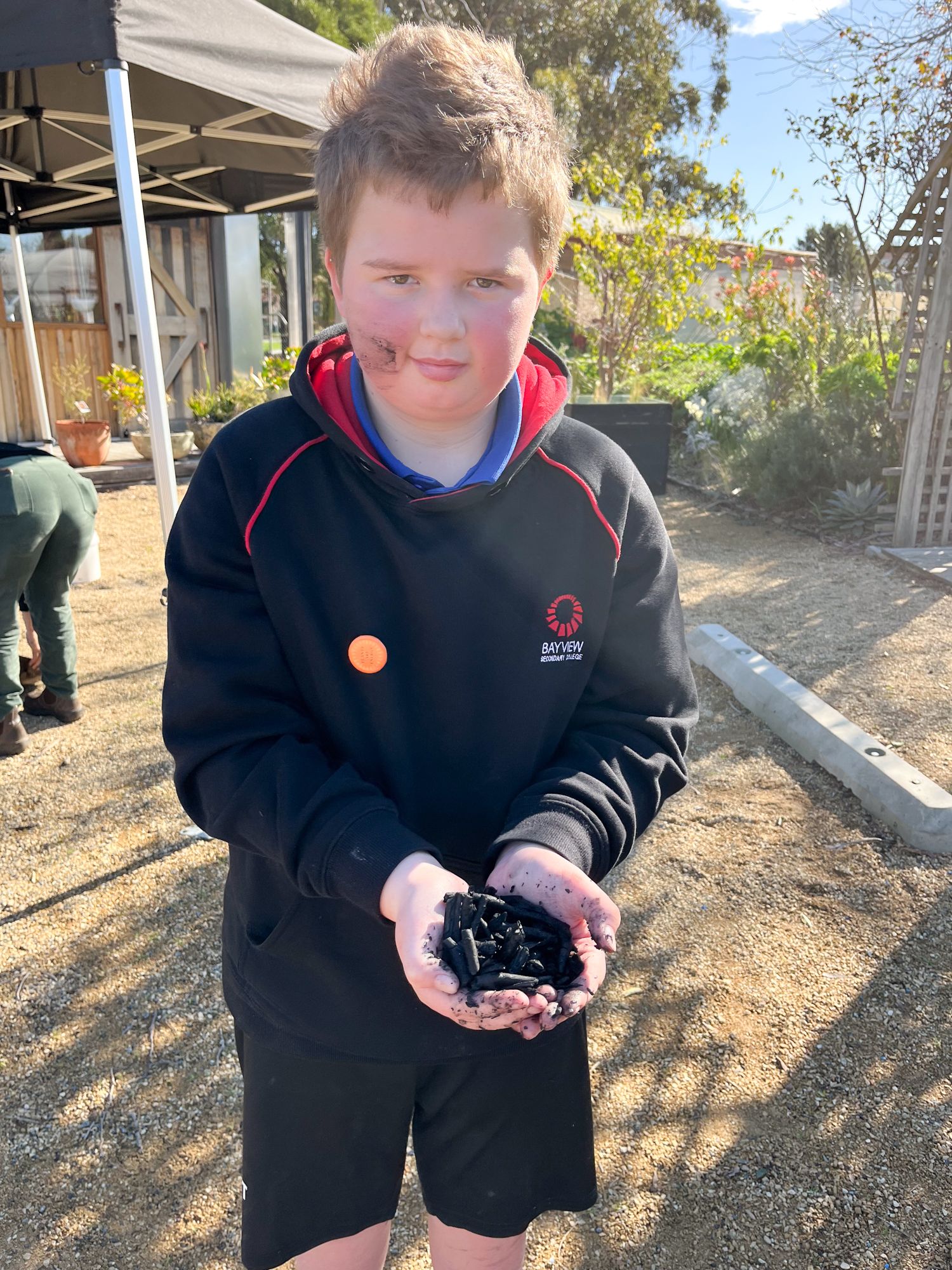 Young boy outdoors, face slightly smeared with dirt, holding black charcoal pieces. He's wearing a black hoodie, looking content under a clear sky.