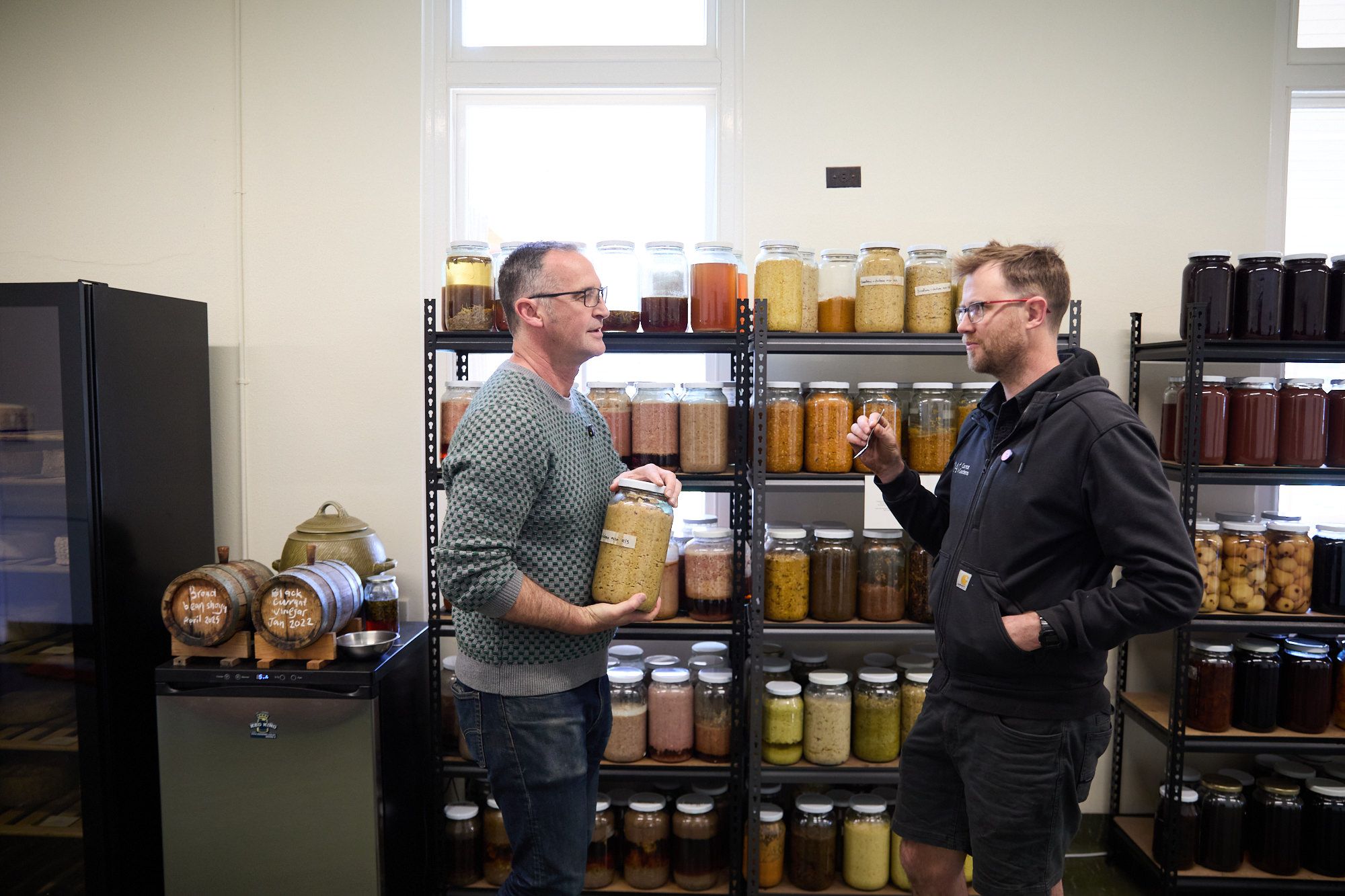 Two men are talking in a room with shelves filled with jars of various preserves. One man holds a jar while the other tastes from a spoon. The atmosphere is casual and engaged.