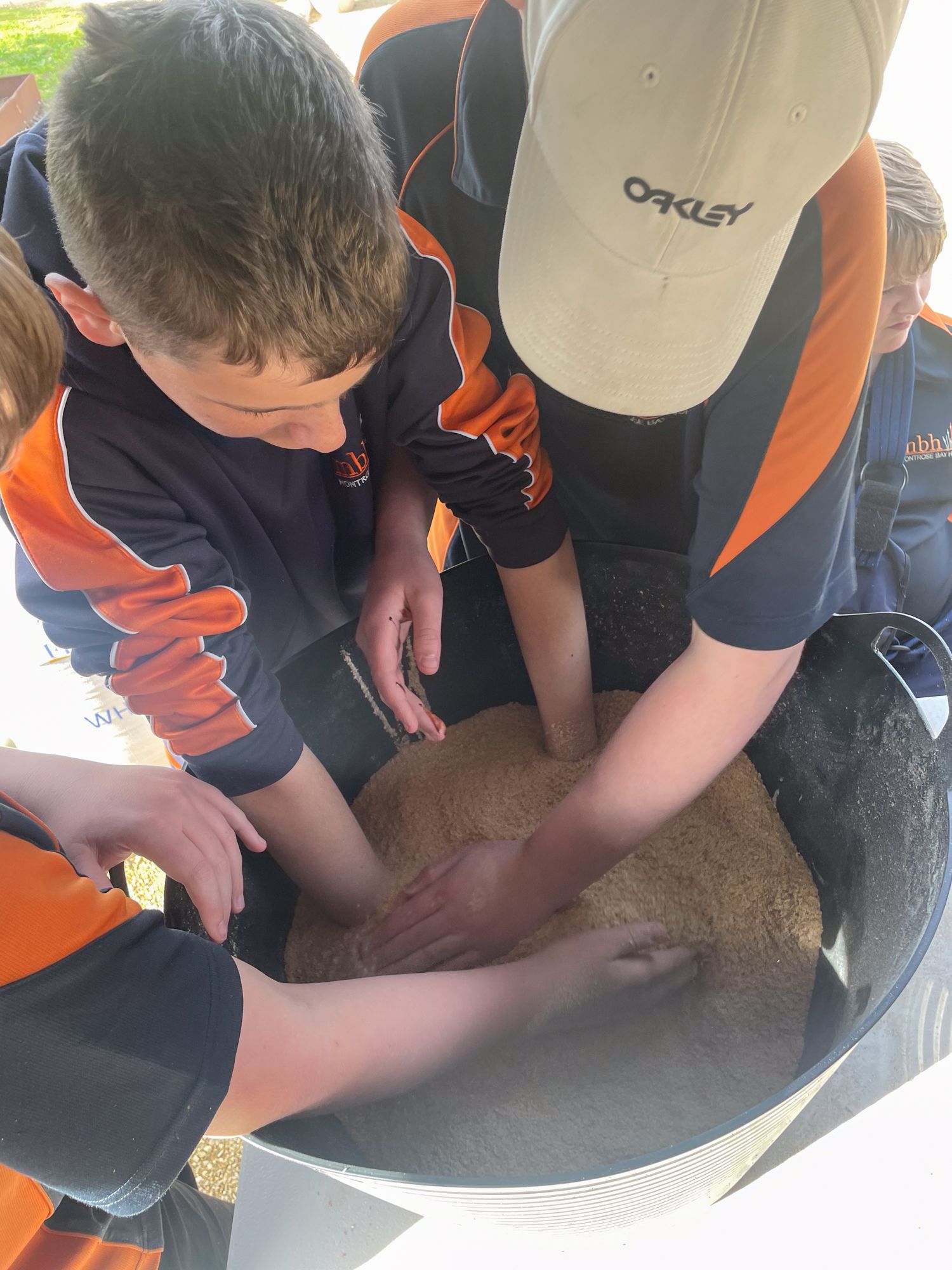Children in uniforms gather around a large container filled with grainy material, possibly sand. They engage with curiosity and teamwork.
