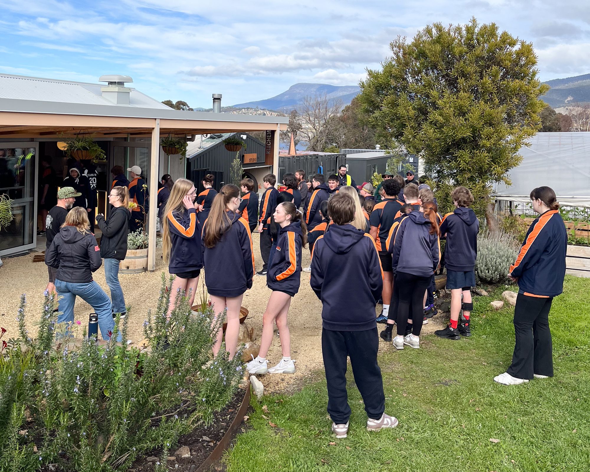 A group of students in navy and orange uniforms gather outside a small building surrounded by greenery. The mood is lively and social.