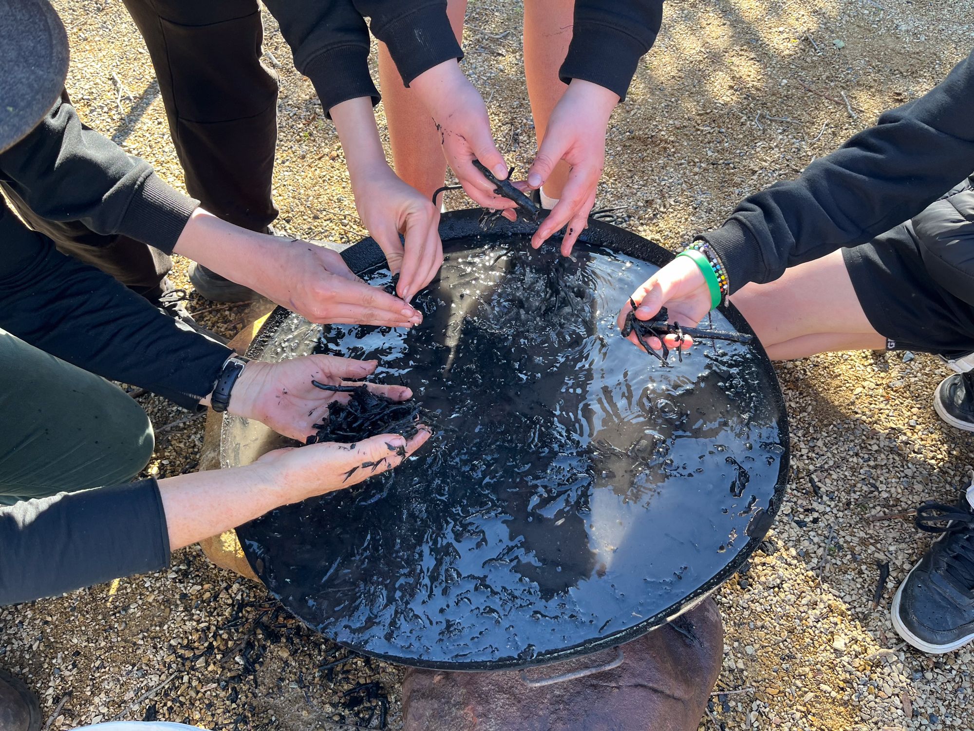 Five hands work together over a large round tray filled with thick, black, tar-like substance outdoors. The focus is on teamwork and texture exploration.