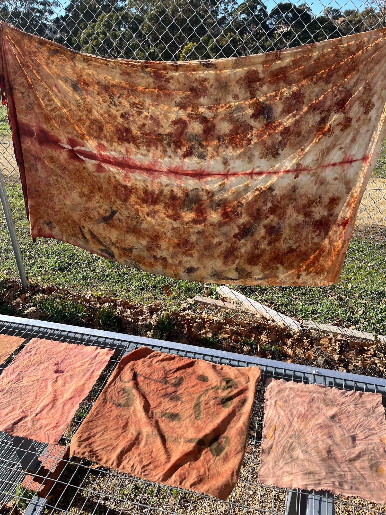 Patchwork of colorful, tie-dyed fabrics dries on a metal rack and a chain-link fence under sunny skies, casting dynamic shadows on green grass.