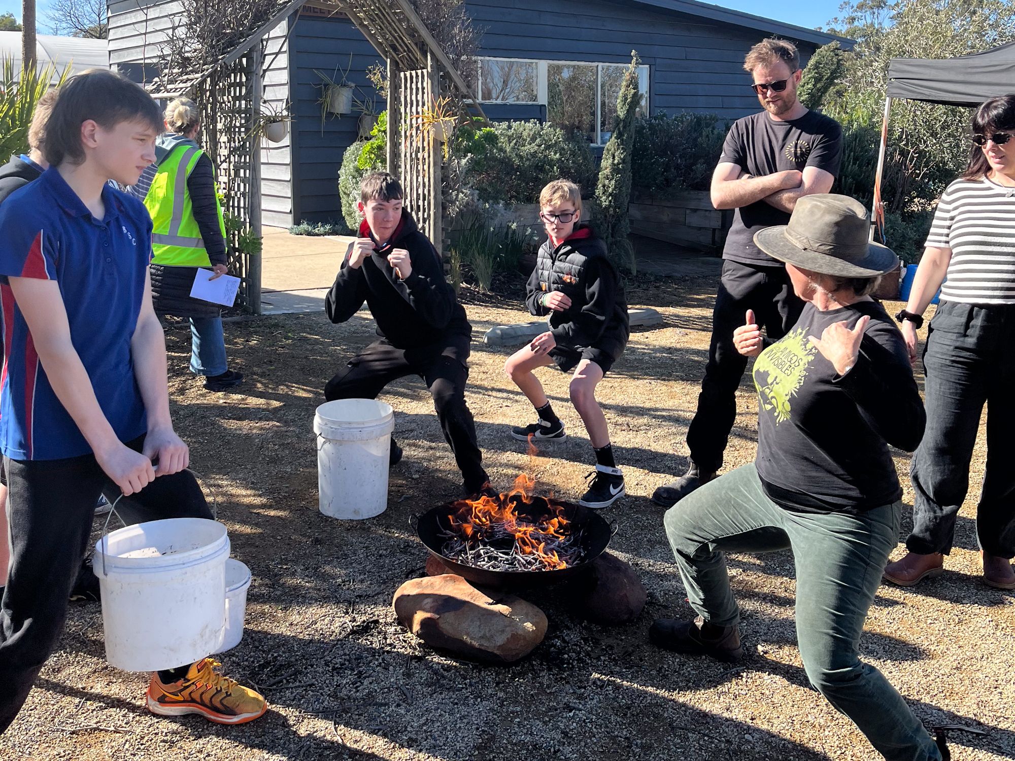 A group of people outdoors, gathered around a fire pit. Two individuals strike playful boxing poses while others watch and smile. The mood is cheerful.