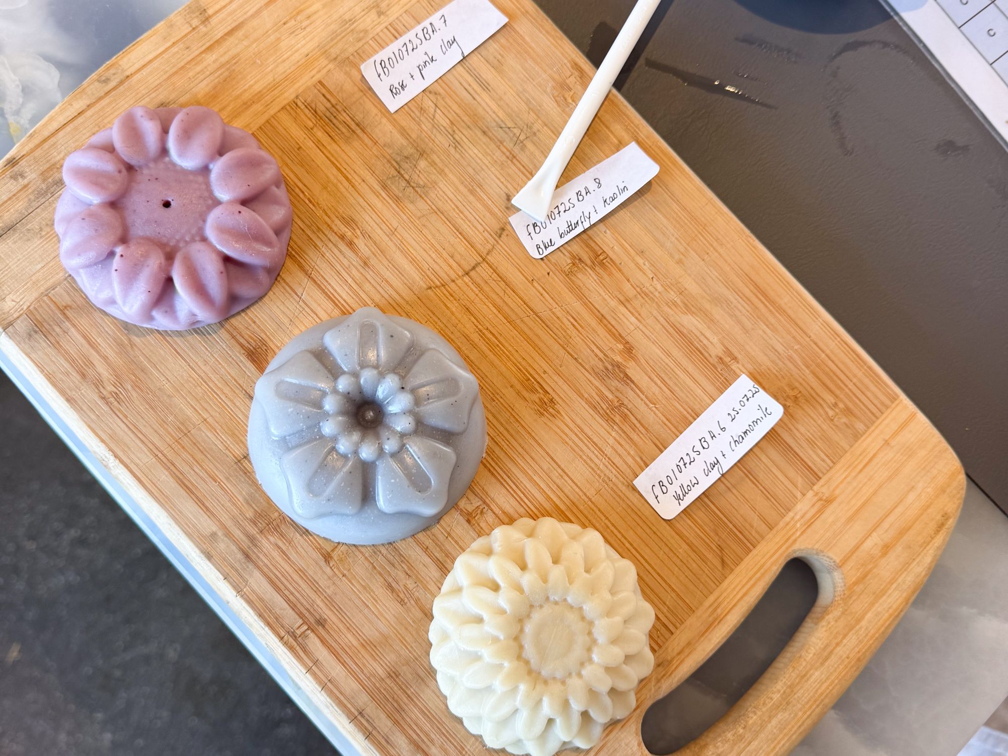 Three intricately molded soaps in pastel purple, gray, and cream on a wooden board. Each soap features floral designs and a small handwritten label.